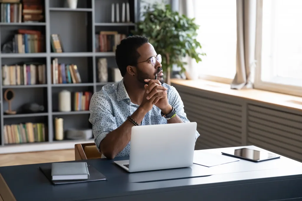 Thoughtful,Multiracial,Businessman,Sitting,At,Table,With,Computer,Looking,Away.